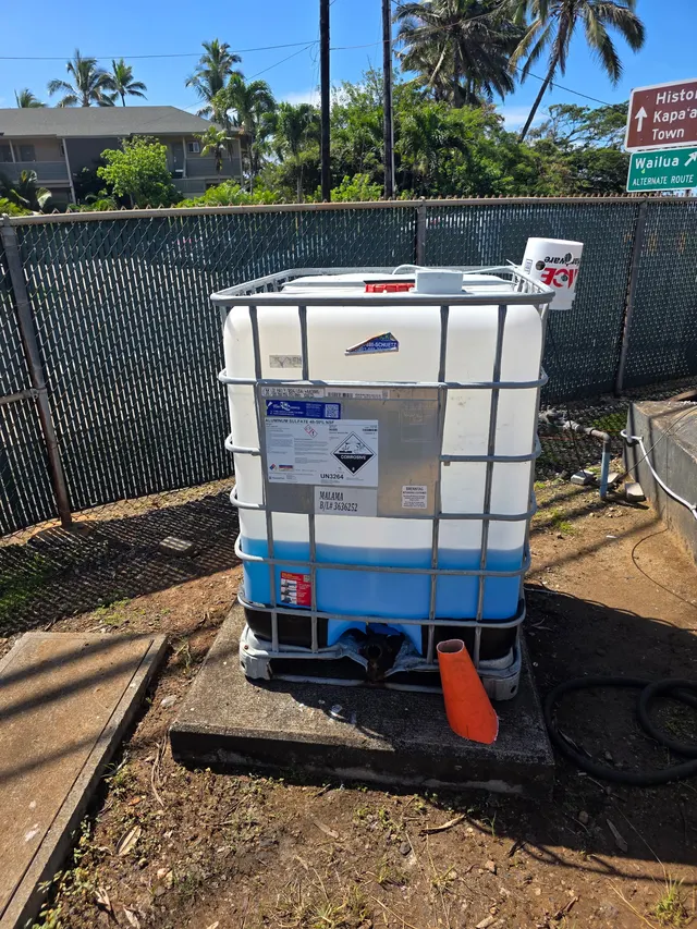 An IBC tote dosing tank in a fenced yard in Kapa'a, Hawaii, with an ultrasonic level sensor on top